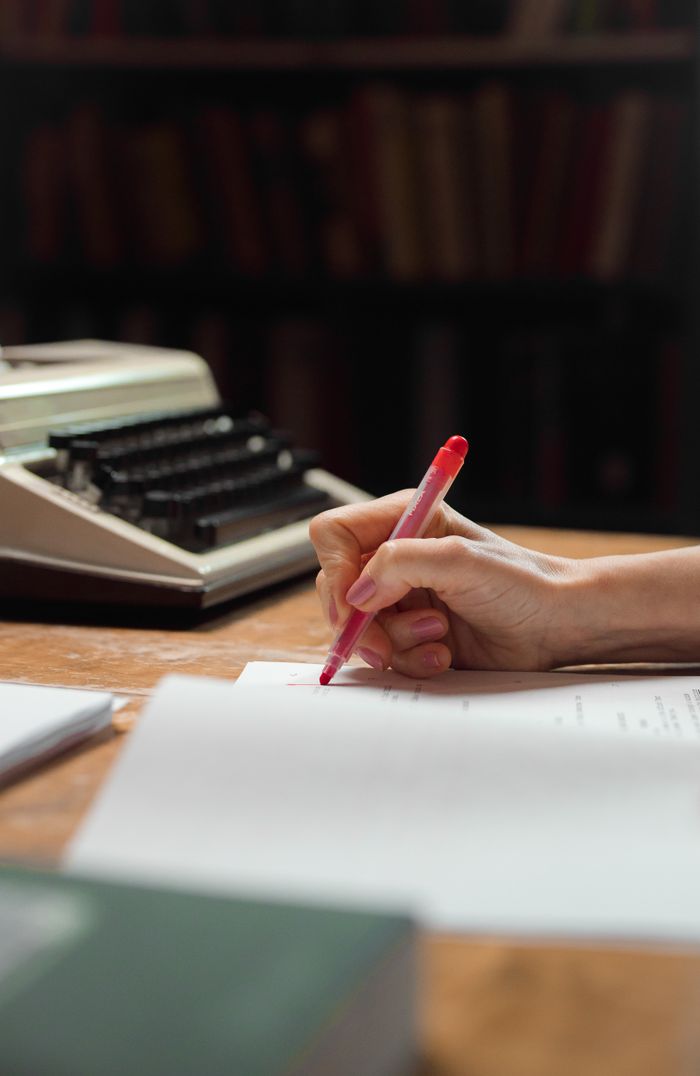 Home A close-up shot of a hand writing on papers with a red pen beside a vintage typewriter.